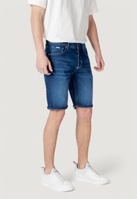 Pantalones cortos de mezclilla en azul oscuro con dobladillos enrollados, que cuentan con cinco bolsillos y una cremallera frontal. Llevados con una camiseta blanca y zapatillas blancas.