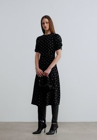 Γυναίκα που φοράει μαύρο μίντι φόρεμα με λευκές πουά, κρατάει μια μικρή μαύρη τσάντα και φοράει μαύρες ψηλοτάκουνες μπότες μέχρι το γόνατο.