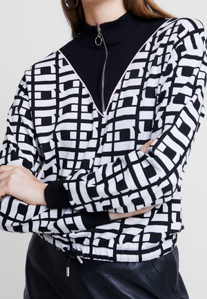 Vrouw die een zwart-wit geometrisch patroon trui draagt over een zwart topje met rits en ringritstrekkertje, armen gekruist, en zwarte broek.