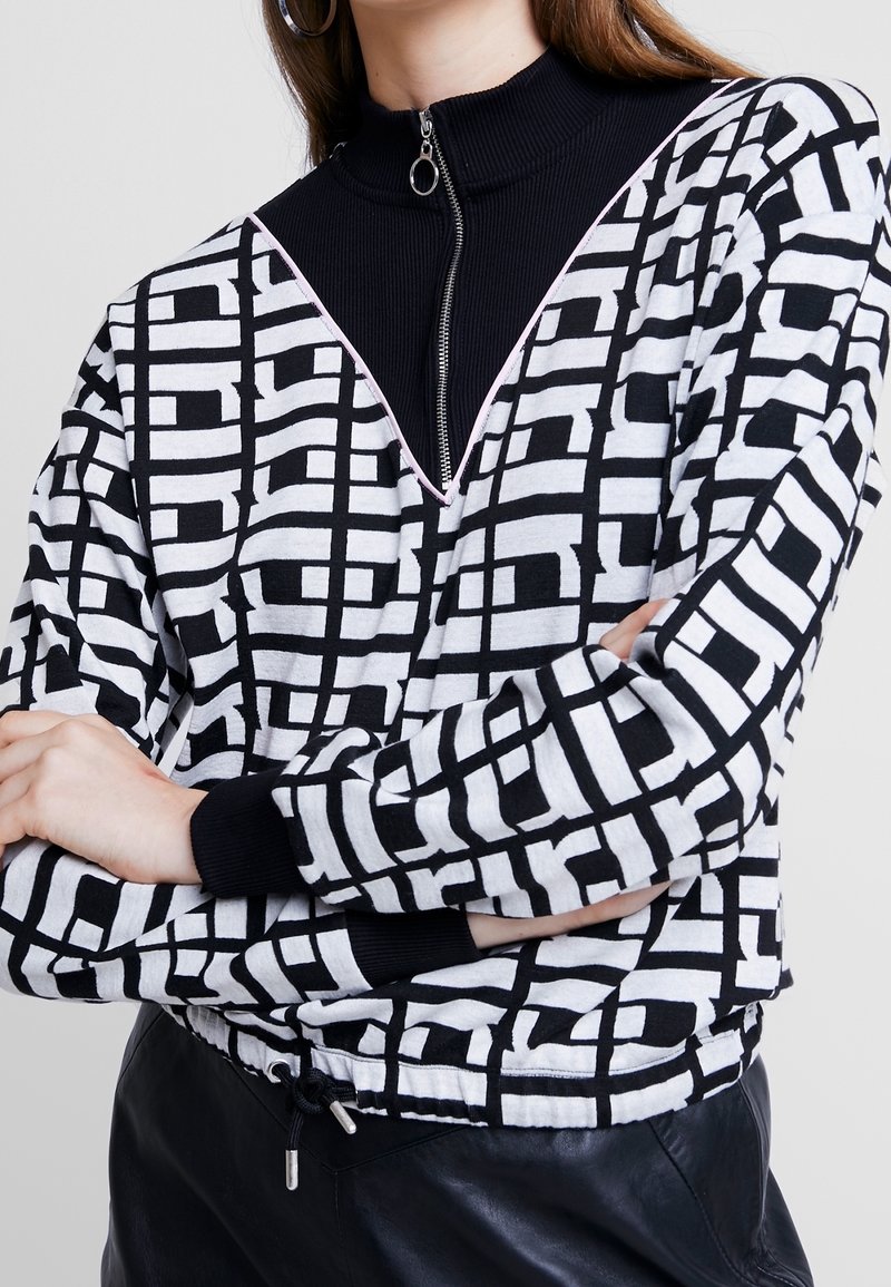 Femme portant un pull à motif géométrique noir et blanc sur un haut noir zippé avec une tirette en anneau, bras croisés, et pantalon noir.