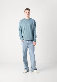 Sudadera azul claro con puños y dobladillo acanalados, combinada con jeans de mezclilla claros. Zapatillas blancas visibles, fondo neutro.