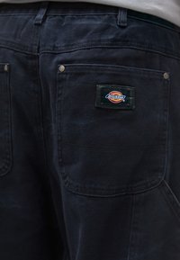 Σκούρα μπλε καμβά παντελόνια εργασίας με υφή, που διαθέτουν δύο πίσω τσέπες και μια μαύρη ετικέτα που εμφανίζει το λογότυπο "Dickies".