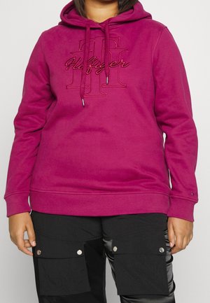 Person iført en magentafarvet hættetrøje med "Hilfiger" logo og sorte cargobukser med knaplommer, stående mod en ensfarvet lys baggrund.