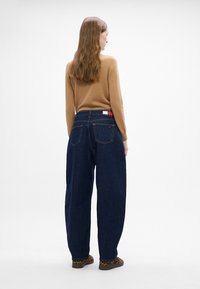 Jeans de mezclilla oscuros de pierna ancha con bolsillos traseros prominentes y una etiqueta de marca. El modelo lleva un suéter beige y zapatos de estampado de leopardo.