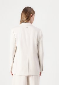 Veste tailleur beige clair avec revers, fermeture à un bouton et fente au dos. Texture lisse et design légèrement structuré.