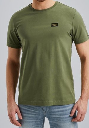 T-shirt en coton vert à manches courtes, col rond, avec une étiquette patch sur la poitrine. Associé à un denim bleu clair. Texture lisse.