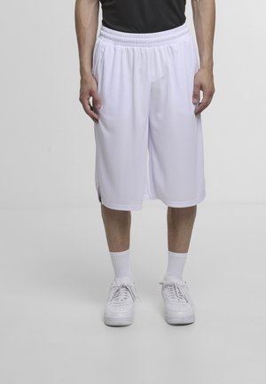 Mężczyzna ubrany w białe, sięgające kolan spodenki do koszykówki, białe skarpety za kostkę i białe trampki, stojący na tle jednolitego jasnoszarego tła.