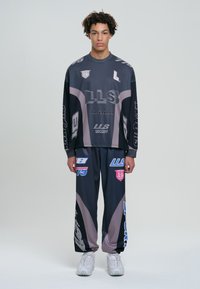Tricou cu mânecă lungă de culoare gri închis, cu logo-uri și grafice îndrăznețe; pantaloni largi asortați, cu accente de gri deschis și culori vii.