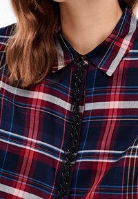 Rutigt skjorta med mönster i röd, marinblå och vit färg, en klassisk krage och dekorativ svart kant längs knappslået.