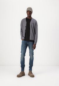 Cardigan gri peste un tricou negru, blugi albaștri și cizme bej. Include o șapcă gri cu logo. Design simplu și croială relaxată.