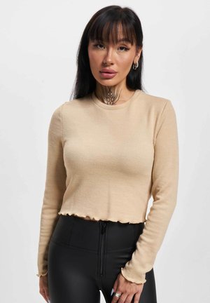 Beige schulterfreies Langarm-Crop-Top mit gewelltem Saum. Enganliegendes Design mit Rundhalsausschnitt und weicher Textur. Kombiniert mit schwarzen High-Waist-Hosen.