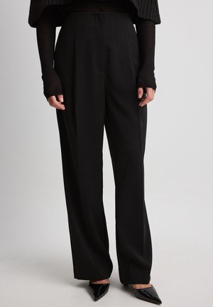 Pantaloni neri su misura con vita alta, pinces e gambe larghe. Il tessuto appare liscio e leggero, abbinato a scarpe nere con punta affilata.
