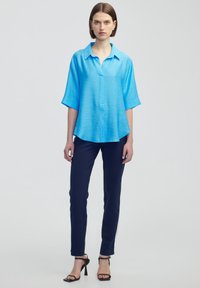 Světle modrá košile s krátkým rukávem a knoflíkovým zapínáním, s límečkem a volnějším střihem, v kombinaci s těsnými námořnickými kalhotami a černými sandály na podpatku.