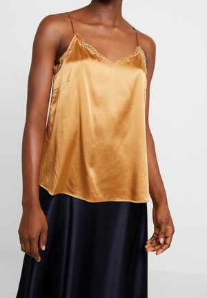 Mujer con una camisola de satén dorado con encaje y tirantes finos, combinada con una falda de satén negra sobre un fondo gris liso.