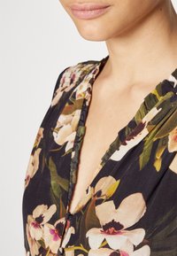 Robe fleurie avec un profond décolleté en V, fond sombre, et des fleurs blanches et roses vives. Comprend une bordure à volants et un détail de bouton discret au niveau de la poitrine.