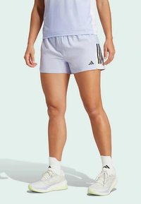 Šviesiai violetiniai sportiniai šortai su juodomis šoninėmis juostelėmis ir Adidas logotipu. Dėvimi su baltais sportbačiais su žaliais akcentais ir baltais kojiniais.