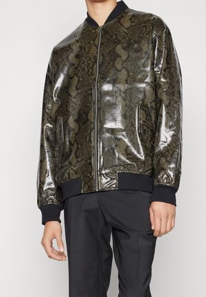 Modelka nosząca błyszczącą bomberkę w ciemnozielonym i czarnym wzorze przypominającym skórę węża, z zamkiem błyskawicznym z przodu oraz czarnymi mankietami i ściągaczem.