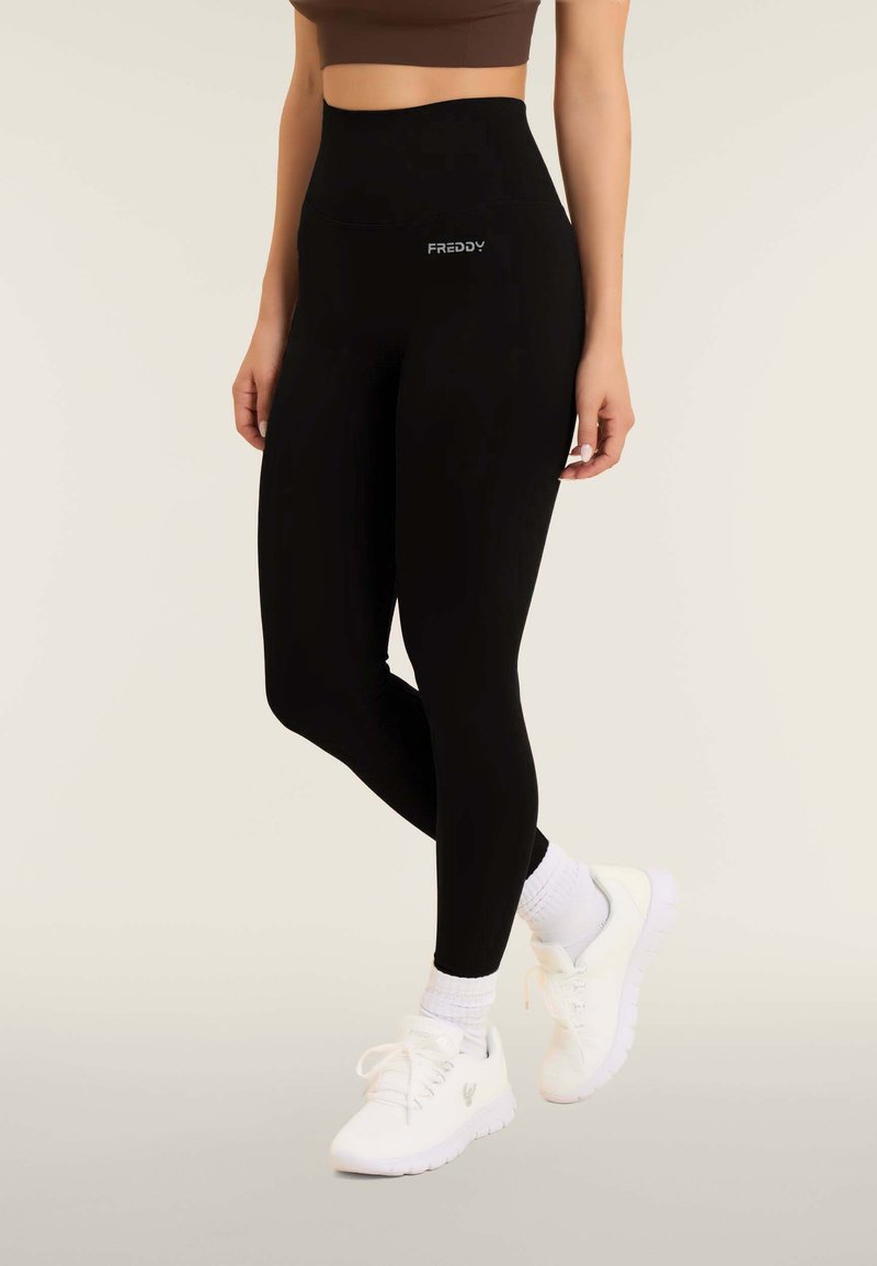 Leggings neri a vita alta con texture liscia e un piccolo logo bianco in vita, abbinati a scarpe da ginnastica bianche e calze.