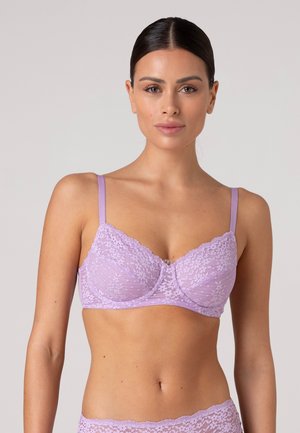 Donna con capelli scuri raccolti indietro che indossa un reggiseno di pizzo lilla e biancheria intima abbinata, rivolta in avanti su uno sfondo chiaro uniforme.