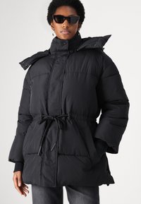 Black padded parka jacket with a high collar, adjustable drawstring waist, and snap buttons. Features large pockets and a hood.
