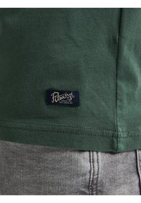 Grünes Baumwoll-T-Shirt mit einem kleinen marineblauen Etikett mit weißem Text, das am unteren Saum eingenäht ist, kombiniert mit grauem Denim-Stoff.