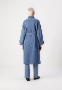 Cappotto in denim con maniche lunghe e vita con cintura, di colore azzurro chiaro. Presenta un colletto, tasche laterali e una vestibilità casual e rilassata.