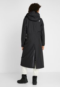 Veste longue noire imperméable avec capuche, poignets élastiques, fente arrière et accents réfléchissants, portée avec des bottes noires et un pantalon blanc.