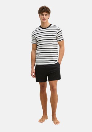 Man met krullend haar, gekleed in een zwart-wit gestreept shirt met korte mouwen en een zwarte korte broek, staand op blote voeten met één hand in zijn zak.