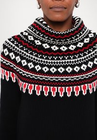 Personne portant un pull noir en tricot avec un empiècement à motifs rouge, blanc et noir et de grandes boucles d'oreilles argentées, visible du cou à la poitrine.