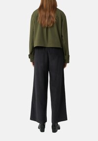 Frau mit langen Haaren, die eine olivgrüne kurze Jacke, weite schwarze Hosen und schwarze Stiefeletten trägt, und mit dem Rücken zu einer einfarbigen Hintergrund steht.