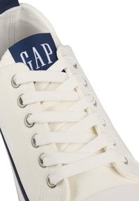 Weiße Leinenschuhe mit einem blauen Textilschild mit "GAP", silbernen Ösen und weißen Schnürsenkeln. Obermaterial aus strukturiertem Stoff und Gummisohle.