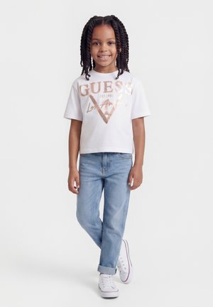 Usměvavé dítě s copánky stojí a má na sobě bílé tričko s nápisem "GUESS Los Angeles", světle modré džíny a bílé tenisky na bílém pozadí.