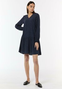 Marineblaues Kleid mit langen Ärmeln; V-Ausschnitt; strukturiertes Muster; ausgestellter Rock; kombiniert mit schwarzen Mules mit einer Kettendekoration.