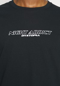 Černé bavlněné tričko s bílým logem "NIGHT ADDICT DYSTOPIA" na přední straně. Kulatý výstřih, krátké rukávy, hladký povrch.