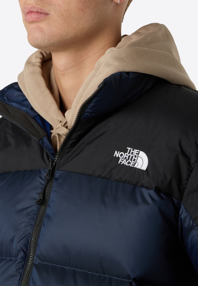 Daunenjacke in Marineblau und Schwarz mit einem Reißverschluss. Merkmale ein weißes Logo auf der Brust und ein beiges Kapuzensweatshirt darunter.