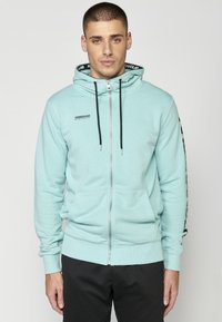 Sudadera con capucha y cremallera en verde azulado claro, con cremallera frontal, dos bolsillos laterales y cordones negros con marca en la capucha y las mangas.