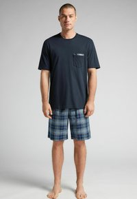 Navyblaues Baumwoll-T-Shirt mit einer Tasche im Karo-Muster, kombiniert mit knielangen Karoshorts in Blau-, Grün- und Grautönen.