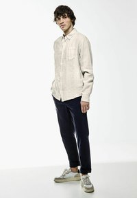 Lichtbeige shirt met lange mouwen van linnen met een borstzak, gecombineerd met donkerblauwe broek en witte sneakers met zilveren accenten.