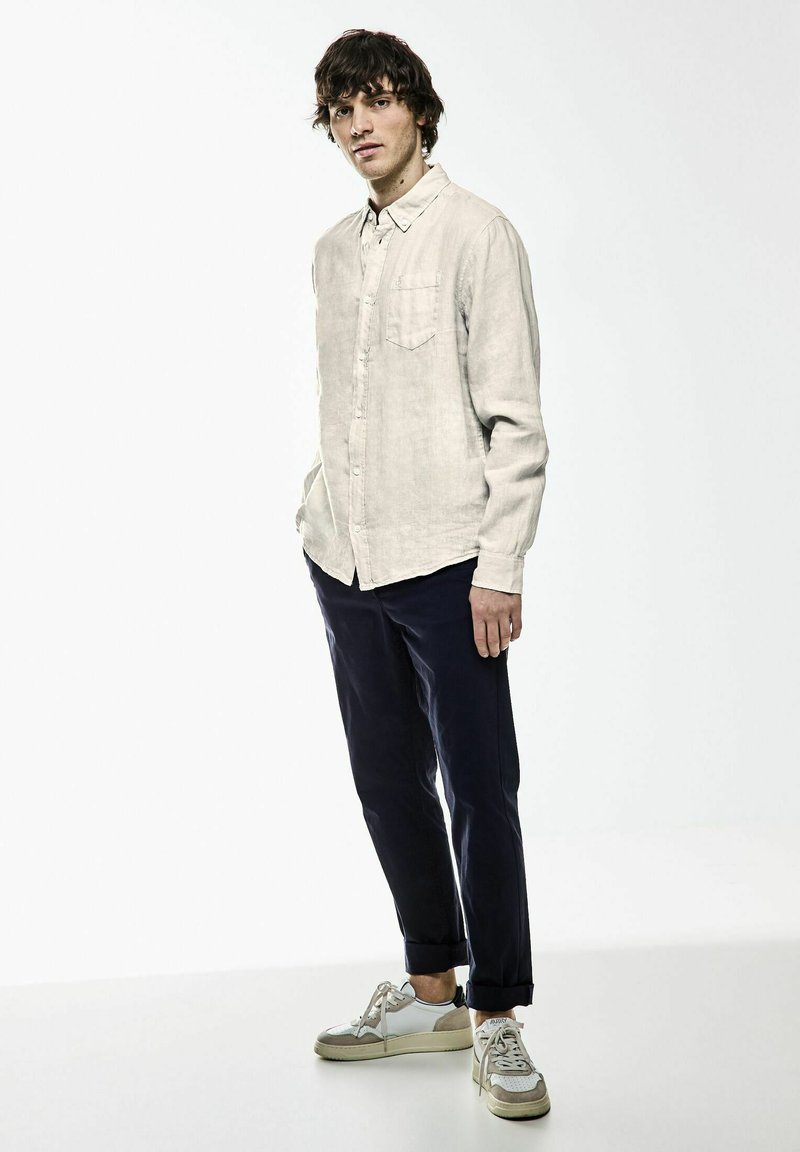 Lichtbeige shirt met lange mouwen van linnen met een borstzak, gecombineerd met donkerblauwe broek en witte sneakers met zilveren accenten.