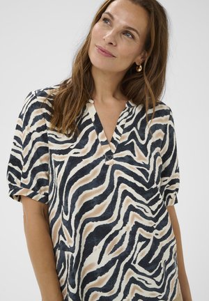 Žena s dugom smeškom kosom nosi zlatne okrugle naušnice i majicu s v-neck izrezom u apstraktnom zebra printu u crnoj, bež i beloj boji.