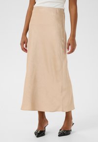Une longue jupe en satin beige au toucher lisse, présentant une silhouette droite et un éclat subtil, associée à des mules noires.