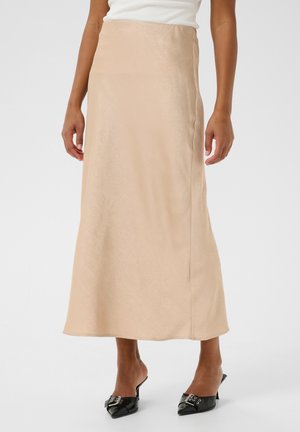 Une longue jupe en satin beige au toucher lisse, présentant une silhouette droite et un éclat subtil, associée à des mules noires.