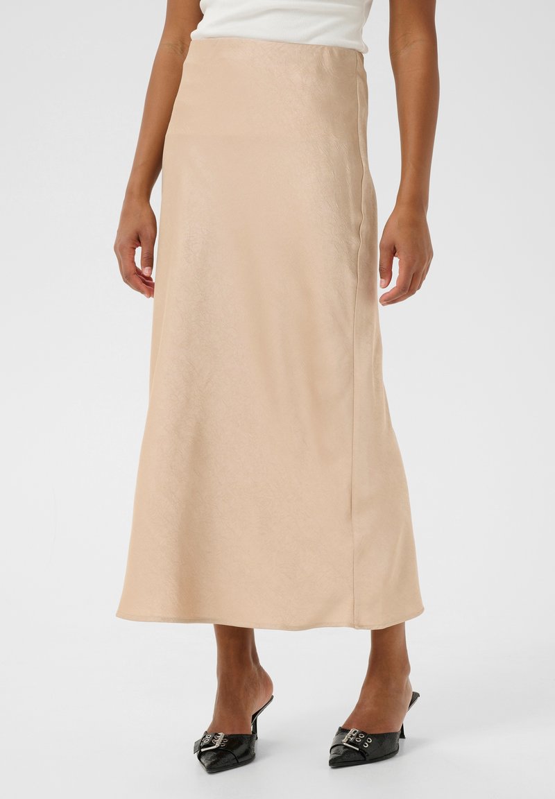 Una larga falda de satén beige con una textura suave, que presenta una silueta recta y un brillo sutil, combinada con unos mules negros.