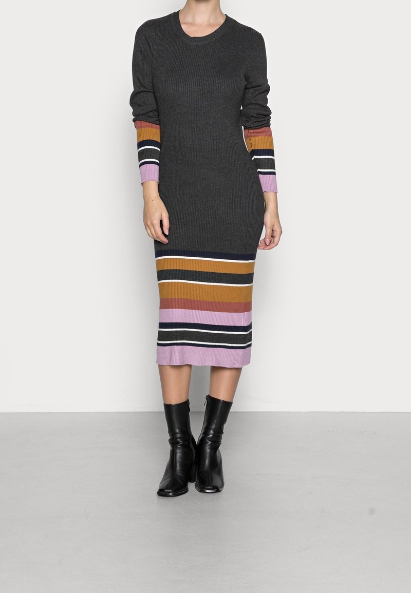 Robe en maille gris foncé avec manches longues côtelées ornées de rayures rose, orange, jaune et bleu marine. Coupe ajustée, longueur genou, portée avec des bottes noires.