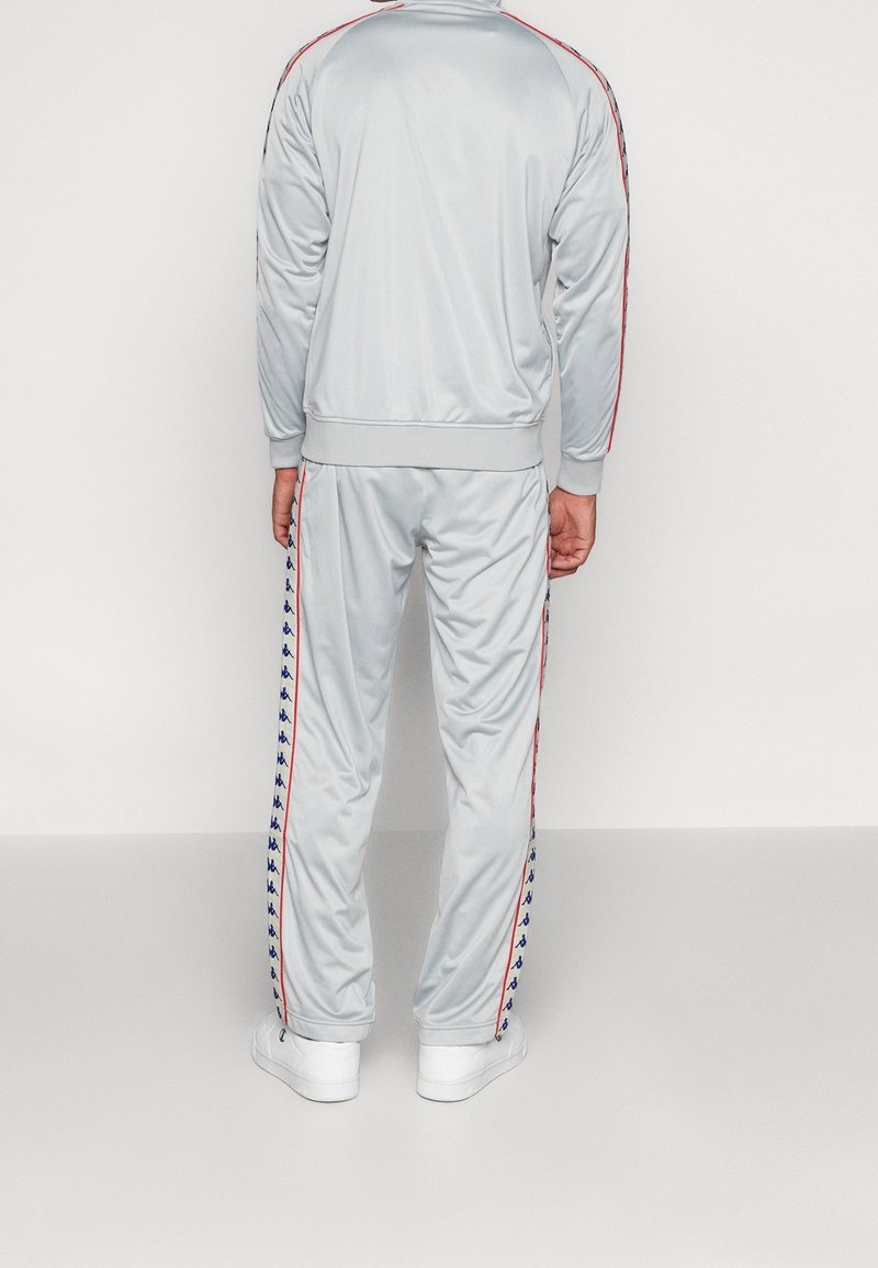 Homme portant un survêtement gris avec des bandes rouges et bleues de logo sur les manches et les jambes, debout face à un mur.