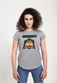 Šedé tričko s krátkým rukávem s motivem Minecraftu, s pixelovanou postavou a výrazným textem v zelených a hnědých akcentem.