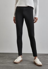 Leggings noires en tissu lisse et extensible ; design taille haute ; associés à des baskets blanches et beige sur un sol gris texturé.
