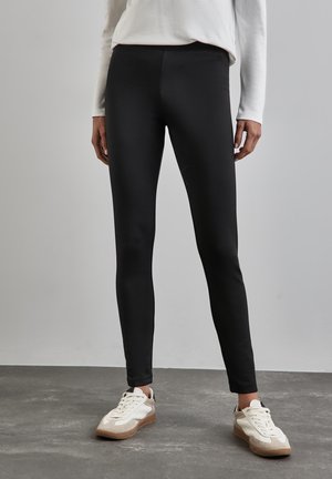 Leggings neri in un tessuto morbido e elasticizzato; design a vita alta; abbinati a sneaker bianche e beige su un pavimento grigio texturizzato.