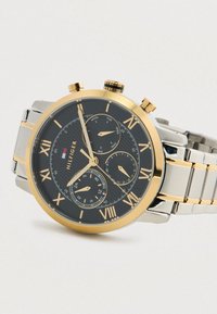 Arany és ezüst karóra fekete számlappal, római számokkal, három segédszámlappal, amelyek mutatják a napot, a dátumot és a 24 órás időt, valamint fém lánc szíjjal.