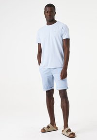 Lichtblauw katoenen T-shirt, gecombineerd met lichtblauwe shorts. Het model draagt bruine sandalen met twee verstelbare banden. Eenvoudig, casual ontwerp.